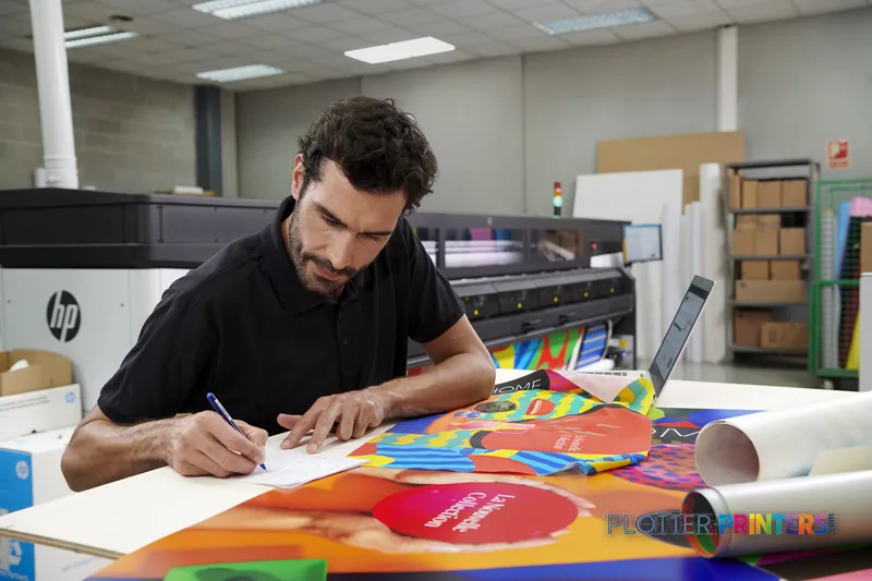 Man reviewing colorful printed materials at workstation with HP Latex 2700 W Printer in background