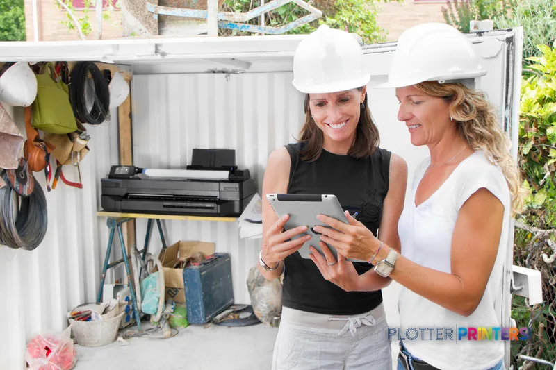 Two women in hard hats reviewing plans on a tablet next to an HP Designjet T120 ePrinter in a job site office