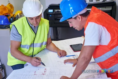 Two construction workers in safety gear reviewing large-format blueprints with HP DesignJet T830 printer in background