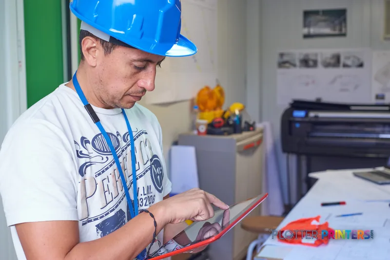 Man in hard hat reviewing printed materials at desk with HP DesignJet T830 printer in background