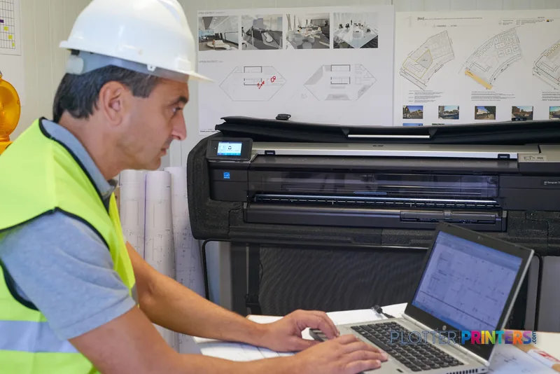 Worker in safety gear operating HP DesignJet T830 24" printer with laptop in architectural office setting