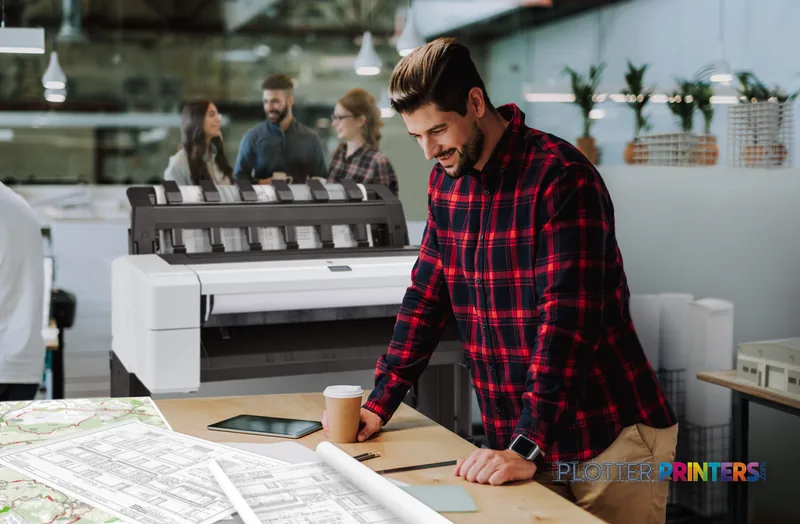 Man in red plaid shirt reviewing large format prints at desk with HP DesignJet T1600 36-in printer in background office sett…