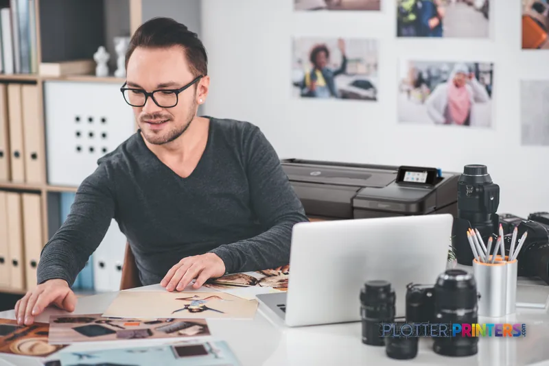 Man working at desk with HP DesignJet Z9+ 24-in PostScript Printer and laptop, reviewing printed photos