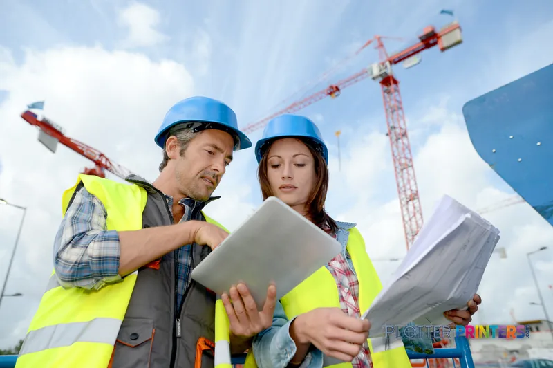 Two construction workers in hard hats and safety vests reviewing large-format prints on a job site with cranes in the backgr…